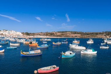 Marsaxlokk Körfezi, Malta 'nın ortasında, sakin mavi sularda düzinelerce parlak luzzu teknesi, beyazlatılmış binalar ve çalışan limanı çerçeveleyen kayalık kıyı şeridi..