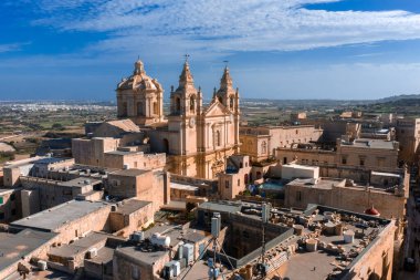 Mdina, Malta 'nın hava manzarasında, St. Pauls Katedrali' nin sıcak kireç taşı ve çıtır çıtır detaylarıyla duvarlı sokaklar, düz çatılar ve parlak öğle saatlerinde tarlalar üzerinde yükseldiği görülüyor..
