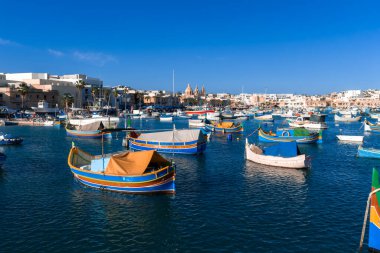 Marsaxlokk Körfezi, Malta, kobalt suyu üzerinde sallanan canlı luzzu tekneleri, şehrin ufuk çizgisi ve gevrek yaz ışığında görünen ikiz çan kuleleri.