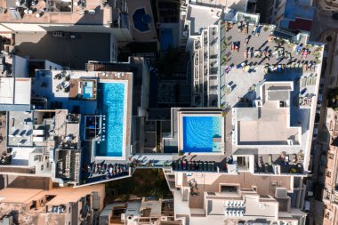 Hava görüntüsü, Malta Mellieha 'daki modern binalarda iki çatı havuzu gösteriyor. Güneşli şezlonglar, gölgeli koltuklar ve parlak gün ışığında teraslarda yaşayan insanlar ve canlı gölgeler..