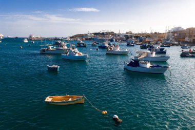 Marsaxlokk, Malta 'nın turkuaz limanında düzinelerce küçük tekne sallanıyor. Önünde sarı bir sandal, geniş panoramik bir manzarayla öğleden sonra ışıkları gövdelerde parlıyor..