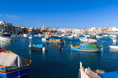 Luzzu tekneleri, Malta 'nın parlak mavi Marsaxlokk limanında balıkçılar demirlerken sallanıyor. Gezinti güvertesi ve kireçtaşı cepheleri gün ortasında ikiz kulelere ve kubbeye yol açar..