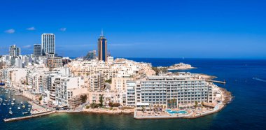 Hava görüntüsü Malta 'daki Sliema yarımadasını, kayalık koylardaki apartman dairelerini ve otelleri, St. Julians' taki Pordomates İş Kulesini, havuzlu, tekneli, açık gün ortası güneşli otelleri gösteriyor..