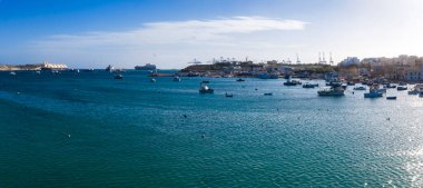 Malta 'nın Marsaxlokk Körfezi' nin panoramik manzarası, turkuaz su, küçük balıkçı tekneleri, büyük gemiler ve parlak gün ışığında eğimli kıyı şeridi boyunca liman vinçleri..