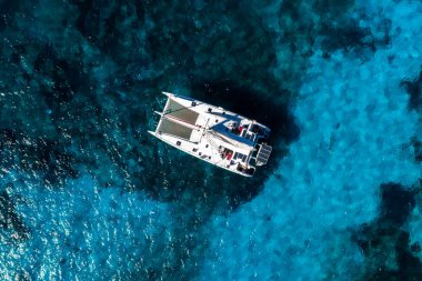 Yukarıdan bakıldığında, Malta, Comino 'daki Blue Lagoon' da demirli beyaz bir katamaran görünüyor. Parlak gün ışığı, berrak dalgalı su, resifler, kum ve teçhizat ayrıntıları görünür..
