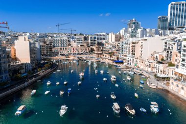 St Julians, Malta 'daki Spinola Körfezi' nin havadan görünüşü, küçük tekneler demirli, kireçtaşı cepheler ve gezinti alanları, modern kuleler ve vinçler öğle güneşi altında