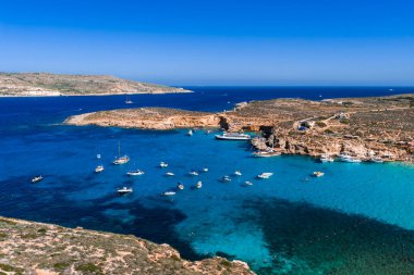Hava görüntülerine göre Mavi Göl, Comino Adası, Malta, turkuaz sığlıklar, kobalt derinlikleri, engebeli kireçtaşı, tekneler ve yatlar, açık yaz ışığı ve görünür deniz yatakları..