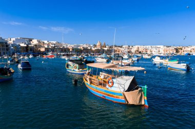 Havadan bakıldığında, Marsaxlokk, Malta 'daki luzzu teknelerinin brandaları, cankurtaranları, deniz kuşlarının daireler çizdiği ve gün ortası aydınlığı altında Parish Kilisesi' nin yükseldiği görülüyor..
