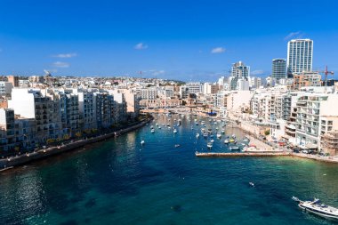 Hava göstergesi, Malta, St. Julians 'taki Spinola Körfezi' ni gösteriyor. Küçük tekneler turkuaz suda yüzer, rıhtımlar kireç taşı blokları ile kıvrılır ve parlak gün ışığında yüksek binalar oluşur..