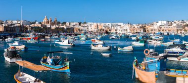 Malta 'daki Marsaxlokk limanı, luzzus ve motorlu tekneleri turkuaz sularda, Pompei Kilisesi' nin Kilisesi 'nin Kireçtaşı Kafeler' in arkasındaki açık gün ışığında gösteriyor..