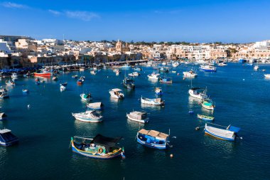 Hava görüntüsü Malta 'da Marsaxlokk Limanı' nı gösteriyor. Turkuaz su, kireçtaşı kasabası, ikiz çan kulesi kilisesi ve gündüz ışığı altında kalabalık bir gezinti alanı..