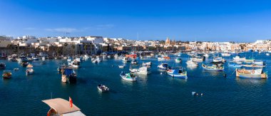 Malta 'nın Turkuaz Marsaxlokk Körfezi' ndeki luzzu teknelerinin parlak panoramik görüntüsü. Pompei Kilisesi kumlu köyün ötesine gün ışığında yükseliyor..