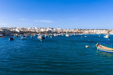Marsaxlokk, Malta gündüz görüşü, luzzu tekneleri parlak mavi sularda sürükleniyor. Kum renkli binalar, gezinti güvertesini berrak bir ışık altında ve sakin bir çalışma havasıyla kaplıyor..