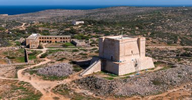 Hava görüntüsü, Comino, Malta 'daki St. Marys Kulesi' ni gösteriyor. Harabeler, tozlu izler ve kayalık çalılıklar var. Derin mavi Akdeniz ufuk çizgisiyle buluşuyor. Uzun gölgeler parlak gün ışığını işaret ediyor.