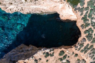 Yukarıdan bakıldığında, Malta, Comino 'daki karenin solunda turkuaz sulara açılan karanlık bir deniz mağarası görülüyor. Güneş yanığı kayalıklar ve çalılıklar öğle vakti uçurumu sarar..
