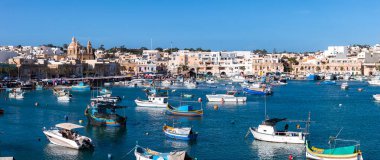 Malta 'daki Marsaxlokk limanı turkuaz su, kireçtaşı kıyısı ve yaz ortasında Pompei Leydisi' nin ikiz çan kilisesi üzerinde renkli luzzu tekneleri gösteriyor..