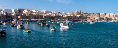 Marsaxlokk Limanı, Malta 'da renkli luzzu tekneleri turkuaz suda yüzer. Kireçtaşı rıhtımı, market tezgahları ve ikiz kilise gündüz aydınlığında yükseliyor..