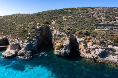 Hava görüntüsü Malta 'daki Comino Adası' nı gösteriyor. Mavi Göl suları kireçtaşı kayalıkları ve küçük bir mağara girişi var. Gün ortası ışığı, temiz su gradyanı, fırça ve teras duvarları..