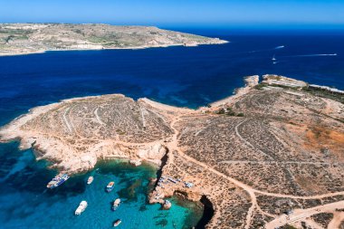 Hava görüntülerine göre Comino Adası, Malta, Mavi Göl turkuvaz suyu, tekneler ve yüzücülerle dar kanallar, kumlu yollar, kanaldan geçen Gozo, parlak gün ortası ışığı.