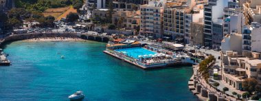 Sliema, Malta 'nın havadan görünüşü Balluta ve Sliema Körfezi' ni gösteriyor, büyük dikdörtgen havuzu, otelleri ve daireleri, küçük bir plajı, güneşlenenleri ve yalnız bir teknesi olan bir lido iskelesi..