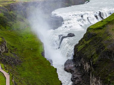 İzlanda 'nın güneybatısındaki Gullfoss, Hvita Nehri' nin iki kat düşüşünün dar bir boğaza düştüğünü, sislerin yükseldiğini, yamaçlarda yürüdüğünü, ziyaretçilerin bulutlu bir ışıkta izlediğini gösteriyor..