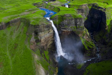 Hava görüntüsü Haifoss 'un İzlanda' nın güneyindeki bazalt kanyona düştüğünü gösteriyor. Bir plato boyunca mavi bir nehir esiyor, sis yükseliyor ve volkanik katmanlar gün ışığında ortaya çıkıyor..