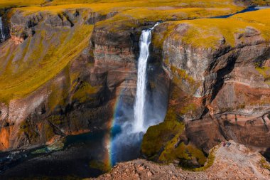 Hava görüntüsü Haifoss 'un soluk bir gökkuşağı, katmanlı volkanik kayalıklar, yosunlu yosun dağları ve parlak gün ışığı altında bir turkuaz havuzu olan bir kanyona düştüğünü gösteriyor..