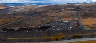 Hava görüntüsü İzlanda dağlık arazilerinde uzak bir alt istasyon gösteriyor. Sütunlar, transformatörler ve şalter kaldırımlı bir yolda oturur ve serin bir ışıkla dolgu boyunca bir kanal akar..