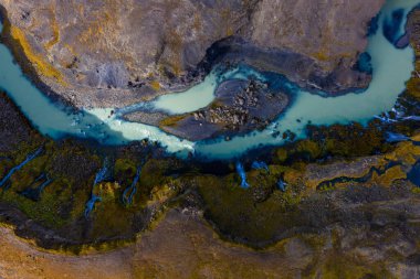 Hava görüntüsü, İzlanda 'daki koyu bazalt ve yosunlu kıyılardan geçen soluk turkuaz bir nehir gösteriyor. Muhtemelen Landmannalaugar' da, yumuşak bulutlu bir ışık altında, mineral renkleri altında..