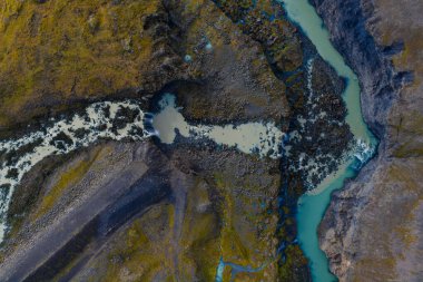 Hava görüntüsü, İzlanda 'da koyu lava ve yosun kesen süt mavisi bir su gösteriyor. Paraşüte doğru daralıyor ve serin, dağınık ışık altında kanyona doğru genişliyor..