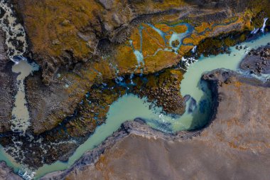 Hava görüntüsü, Landmannalaugar yakınlarındaki İzlanda 'da koyu lav ve yosun üzerine örülmüş sütlü bir turkuaz nehir gösteriyor. Yumuşak yayılmış ışık, çağlayanları ve dalgalı çökeltileri ortaya çıkarır..