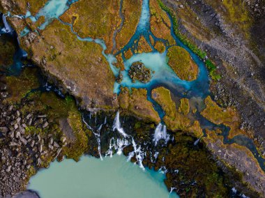 Hava görüntüsü lav ve bazalt üzerinde sütlü turkuaz buzul akıntılarının Sigoldugljufur yakınlarındaki İzlanda dağlarında küçük bir şelale ve mineral havuzuna dönüştüğünü gösteriyor..