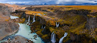 Hava görüntüsü İzlanda 'daki Sigoldugljufur Kanyonu' nu gösteriyor. Bazalt kayalıklarından, kara kara düşünen bulutların altındaki sütlü buzul nehrine dar şelaleler düşer..