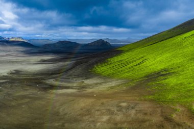 İzlanda 'da Landmannalaugar yakınlarında koyu kül ve lav tarlalarıyla kaplanmış bir yosun örtüsü. Tepeler gibi uzak tepeler ve kraterler yumuşak ışıkla ağır bulutların altında otururlar..