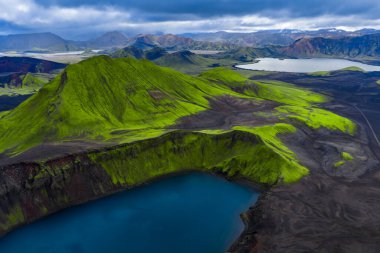 Havadan bakıldığında İzlanda, Landmannalaugar 'da derin mavi bir krater gölü yakınında volkanik bir yosun görünüyor. Dağınık gün ışığı, katmanlı lav kumu ve ryolit zirvelerini ortaya çıkarır..