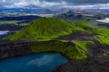 Karanlık lav sahalarının üzerinde yosunlu volkanik bir tepenin havadan görünüşü, İzlanda 'daki üssünde Landmannalaugar yakınlarındaki mavi bir krater gölü, kara kara kara düşünen bulutlar ve dağınık ışık saçan bir göl..
