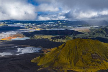 Hava görüntüsü, alçak bulutlar ve serin ışık altında Landmannalaugar yakınlarındaki İzlanda Highlands 'ta yosun yeşil volkanik koni, siyah lav alanları ve çok renkli rhyolit çıkıntıları gösteriyor..