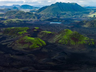 Hava görüntüsü, İzlanda Highlands 'taki engebeli rhyolite dağları ile mavi bir göl yakınındaki siyah lav üzerinde yosunlu kül konileri gösteriyor. Yumuşak gün ışığı ve bozuk bulutlar kontrastı ekler.