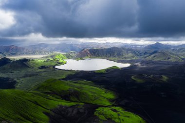 İzlanda 'da engebeli dağların ve lav tarlalarının ortasındaki oval krater gölü. Fırtına bulutları arasından geçen güneş ışınları yosunlu ryolite tepelerini aydınlatıyor..