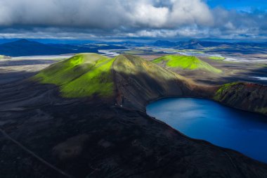 Havadan bakıldığında İzlanda, Landmannalaugar 'daki derin mavi krater gölü yakınlarında yosun yeşili rhyolite tepeleri görülüyor. Güneş ışığı alçak bulutları kırıyor, siyah lav ve kül ovalarını vurguluyor.