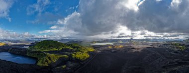 Landmannalaugar yakınlarındaki İzlanda volkanik dağlarının hava manzarası, yosun yeşili rhyolite tepeleri, siyah lav alanları ve hızlı hareket eden bulutların altında mavi bir krater gölü..