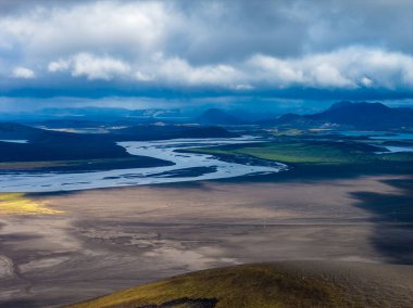 Hava görüntüsü, İzlanda 'nın dağlık bölgelerindeki karanlık volkanik ovalarda örülmüş buzul nehirleri, yosunlu araziler ve öğle vakti ışık bacalarının altında uzak rhyolite dağları gösteriyor..