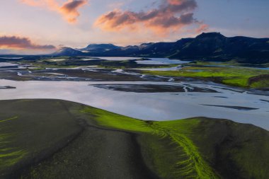 Güneş batımında İzlanda 'da karanlık volkanik kumları aşan örülmüş buzulların hava manzarası, yeşil yosun yamaları ve engebeli dağlar ve tepeler..
