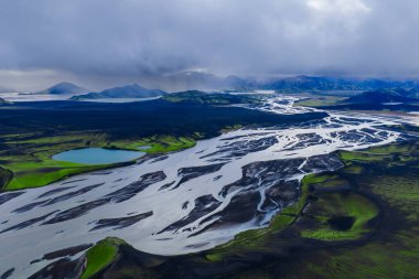 Landmannalaugar yakınlarındaki İzlanda dağlık arazilerindeki siyah kumlar ve yosun tarlaları boyunca örülmüş buzul nehrinin havadan görünüşü, turkuaz göl ve bulutlu ışık kontrastı.