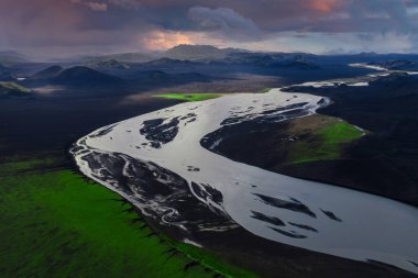 Hava görüntüsü, İzlanda 'nın Landmannalaugar yakınlarındaki siyah bir volkanik ovayı geçen örülmüş bir buzul nehri gösteriyor. Uzak dağların üzerinde yosun yamaları ve alacakaranlık bulutları var..
