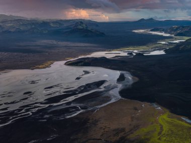 Örülmüş buzul nehirleri, İzlanda 'da Landmannalaugar yakınlarında siyah volkanik kumlar ve yosun arazileri boyunca esiyor. Düşük akşam ışığı ve fırtına bulutları rhyolite zirvelerini gösteriyor.