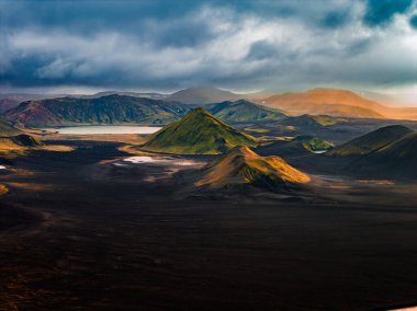 Landmannalaugar yakınlarındaki İzlanda volkanik dağlarının havadan görünüşüne göre tuff konileri, siyah lav kumları, soluk bir göl, yosun yamaları ve gün sonunda bulutların arasından güneş ışınları görünüyor..
