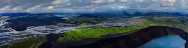 Landmannalaugar veya Fjallabak 'daki İzlanda dağlık arazilerinin hava manzarası, siyah kumlar üzerinde örülmüş buzul nehirleri, yeşil yosunlu bir plato ve kırık bulutların altında turkuaz bir krater gölü gösteriyor..