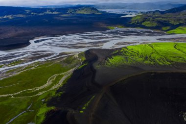 İzlanda 'daki siyah kum ve yosun tepeleri boyunca örülmüş nehirlerin hava manzarası, muhtemelen Landmannalaugar veya Fjallabak, yumuşak ışık altında ryolit yamaçlar ve alçak bulutlar altında.