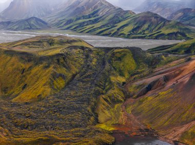 İzlanda 'daki Landmannalaugar' ın hava manzarası çok renkli rhyolite tepeleri, örülmüş nehir ovaları ve yumuşak bulutlu ışık altında yosunlu yosunlu yosunlu yamaçlarda koyu bir lav akışı gösteriyor..